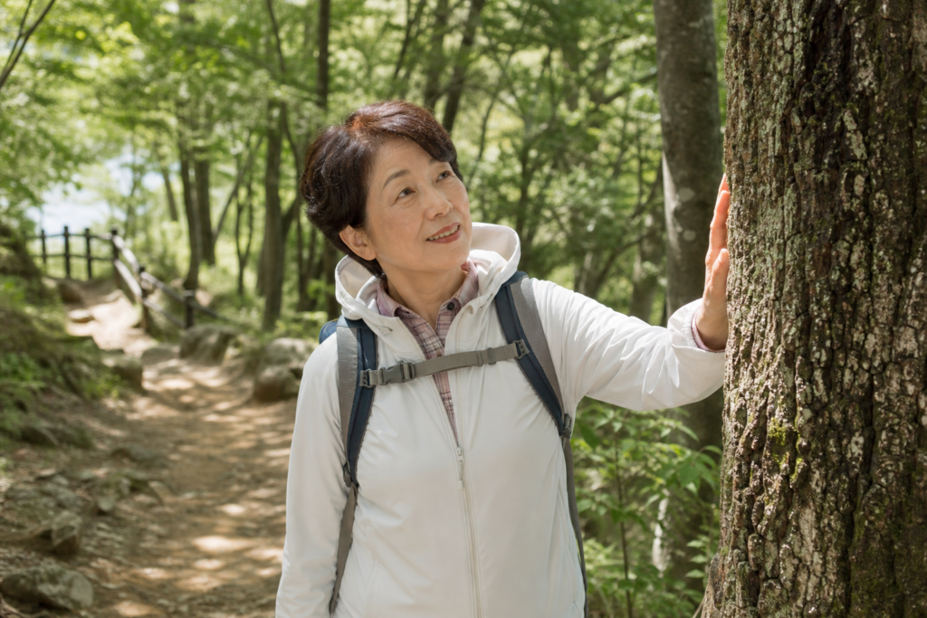 森の小道を歩きながら木に触れて微笑む60代の日本人女性、あたたかな木漏れ日の中の情景
